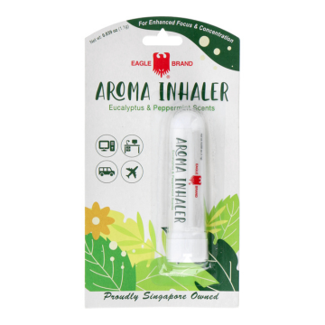 The image shows the packaging of the Eagle Brand Aroma Inhaler with the brand's red eagle logo at the top. The product name, "Aroma Inhaler," is in green, followed by "Eucalyptus & Peppermint Scents." Icons indicate it's suitable for use while traveling, at home, or on the go. The bottom features floral designs and the tagline "Proudly Singapore Owned." The inhaler stick is displayed next to the text.