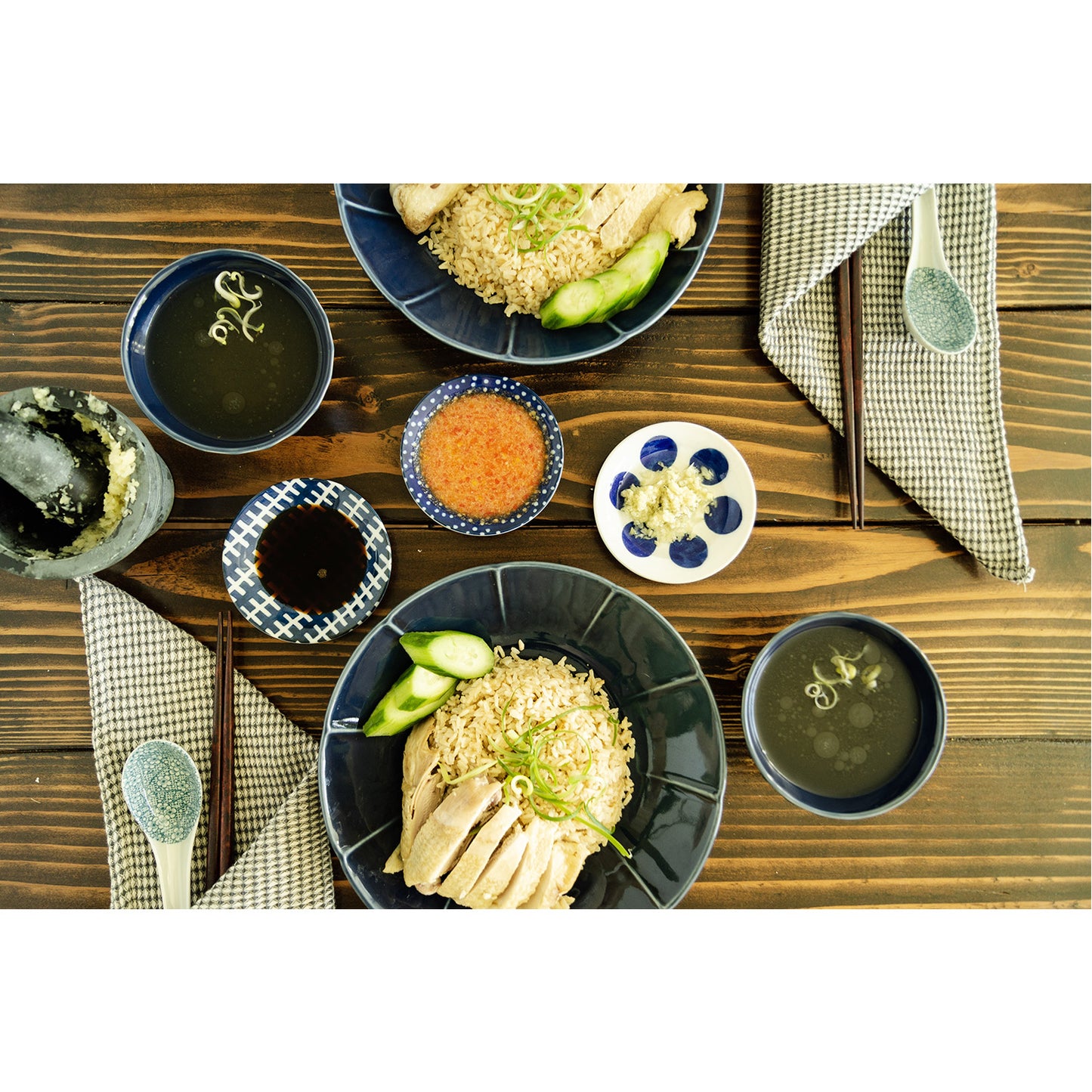 Lifestyle photograph of Hainanese sachets. A bowl of hainan rice and steamed chicken with chopsticks, variety of sauces, mortar, pestle, and broth is pictured.